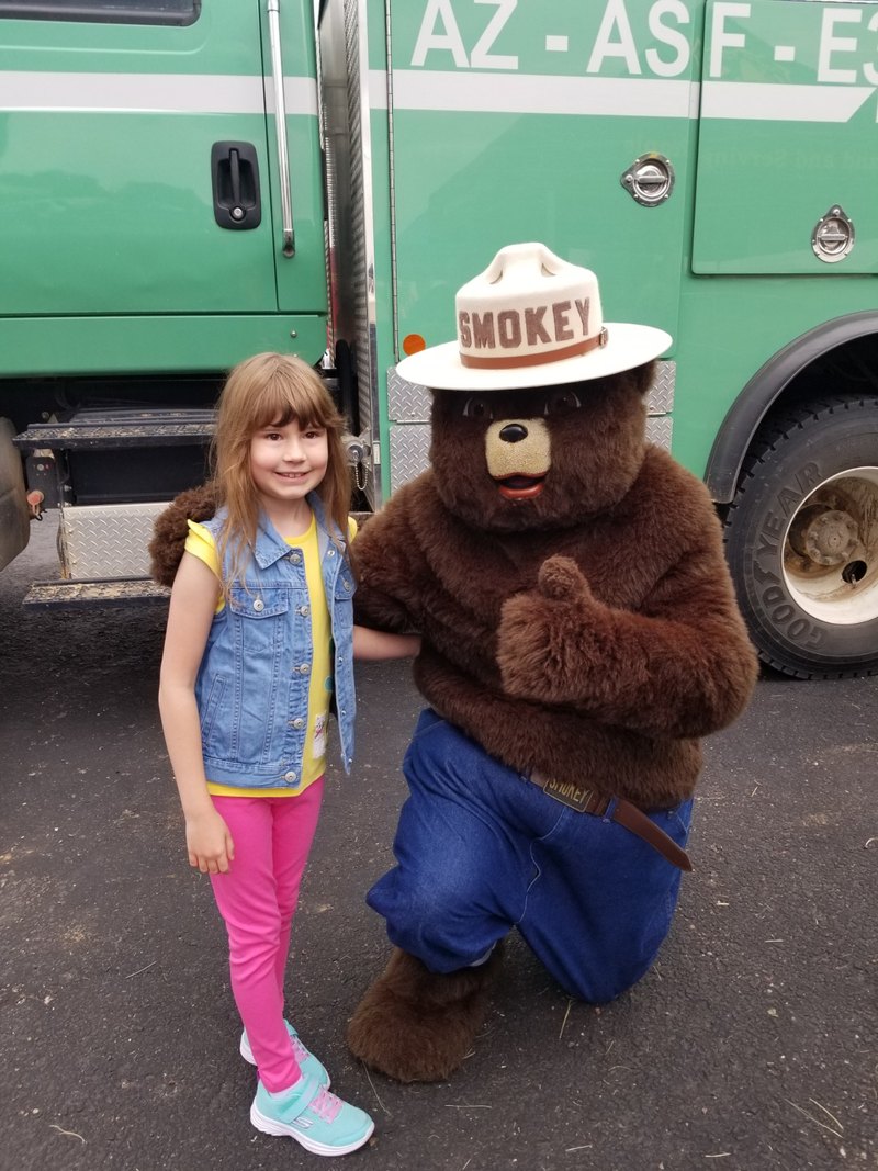 Smokey Bear kneels beside a young visitor in front of a green Forest Service vehicle